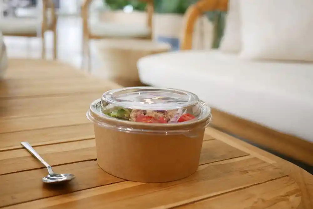 Tahta masa üzerinde, yanında kaşık bulunan şeffaf kapaklı bir kapta salata