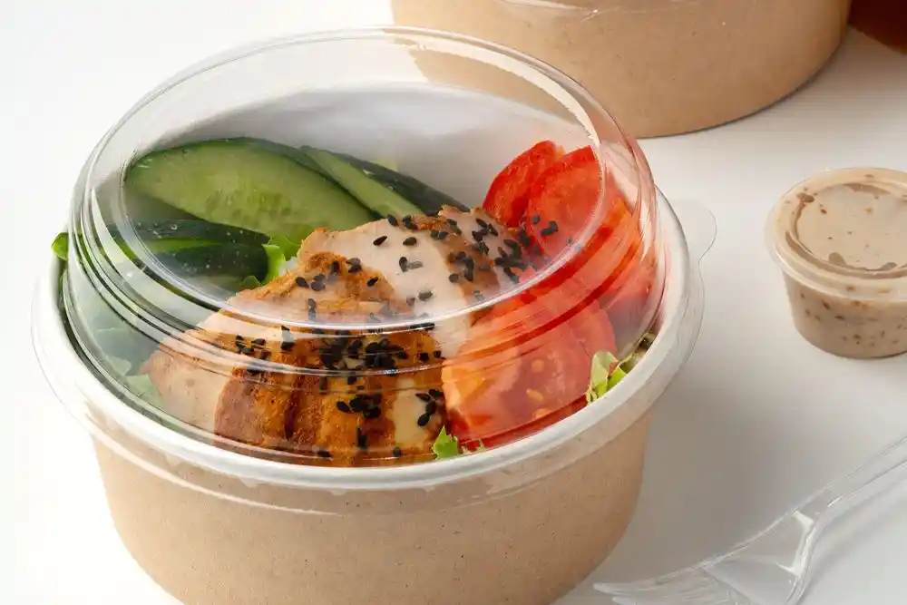 Şeffaf kapaklı kapta tavuk, salatalık, domates ve yeşilliklerden oluşan salata; yanında sos kabı