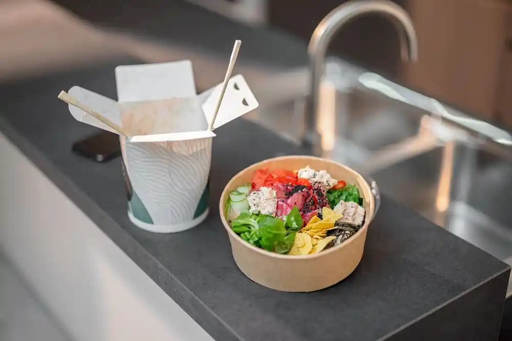 Siyah tezgâh üzerinde çeşitli malzemelerle hazırlanmış bir salata kasesi ve  karton kapak paket servis bardağı